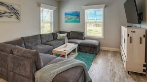 A living room with a grey sectional sofa, a small coffee table, and a television on a cabinet. There are two windows with blinds, a ceiling fan, and a painting on the wall. - New! Oceanview Oasis whole house - Theater - Screened Porch - Sundeck - Grill (Marathon)
