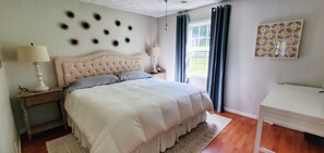 Un dormitorio con cama y cabecera capitonada, dos mesitas de noche con lámparas, una ventana con cortinas y un elemento decorativo en la pared.