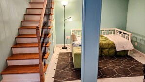A bedroom with a wooden staircase, a bed with a green blanket, a floor lamp, and a patterned rug.