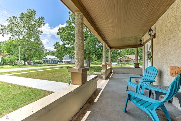 A covered porch with two blue chairs and a table, a concrete pathway, and a grassy area with trees and houses in the background. - Beit Shalom (House of Peace) (Siloam Springs)