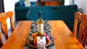 Une table à manger agrémentée d’un chemin de table à motifs floraux, une bouteille d’huile d’olive et un panier de fruits. Un canapé turquoise et un téléviseur se trouvent à l’arrière-plan.