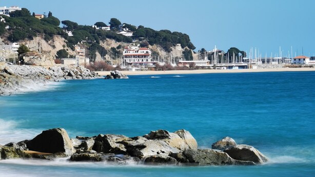 Une plage à l’eau d’un bleu limpide, avec des rochers et des bâtiments situés sur une colline en arrière-plan.