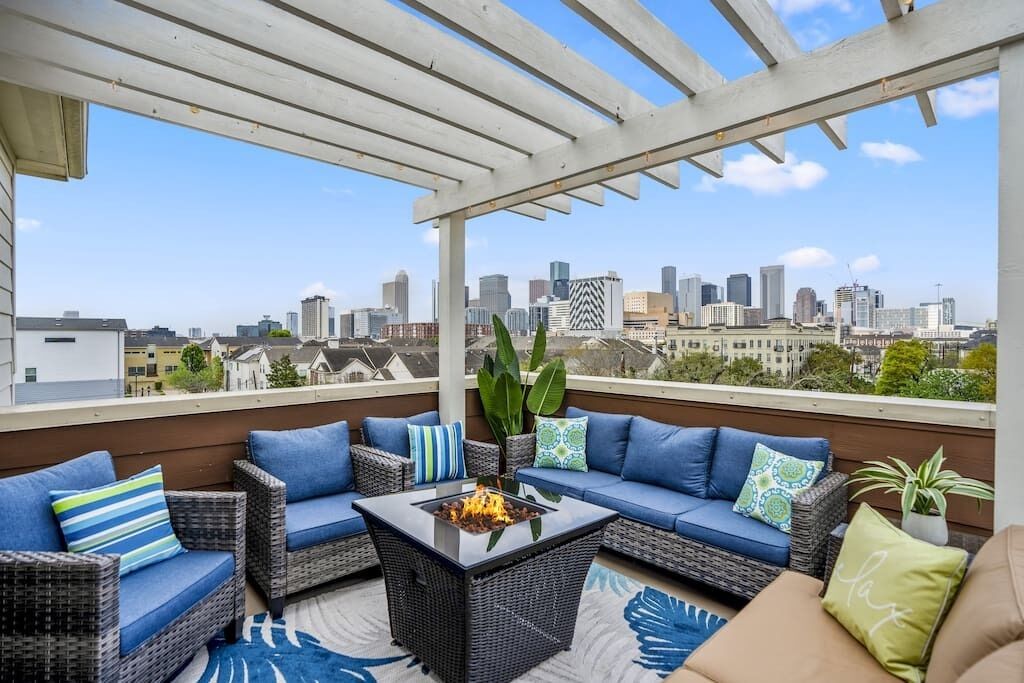 A rooftop patio with wicker furniture, a glass-top table with a fire feature, and a city skyline view.