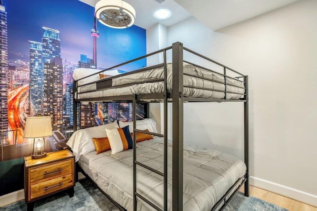 A bunk bed room with a cityscape mural, a bedside table with a lamp, and a nightstand with drawers.