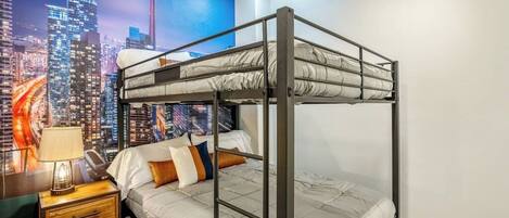 A bunk bed room with a cityscape mural, a bedside table with a lamp, and a nightstand with drawers.