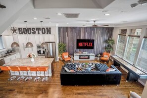 A modern kitchen with a bar area, a living room with a black sofa and orange chairs, a TV displaying 'NETFLIX,' and a 'HOUSTON' sign on the wall.