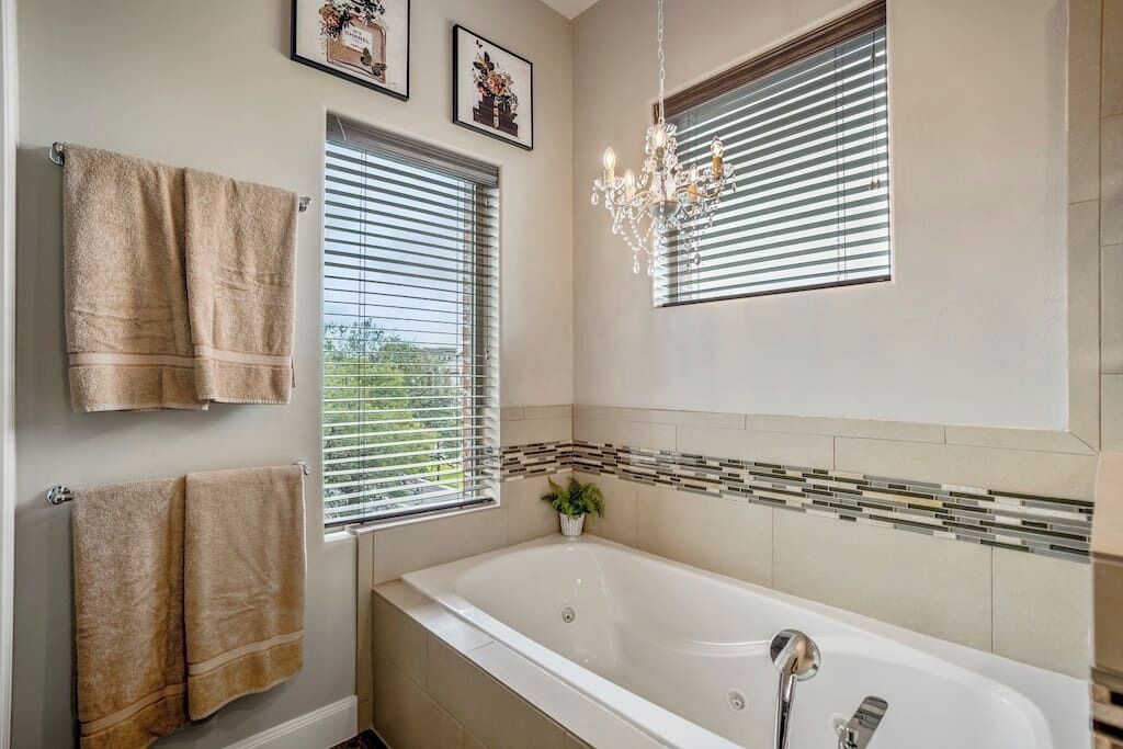 A bathroom with a bathtub, two towels, a chandelier, and a window with blinds.