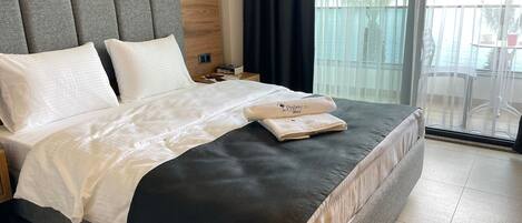 Habitación de hotel con una cama grande, cabecera gris, ropa de cama blanca y vistas al exterior a través de un ventanal.