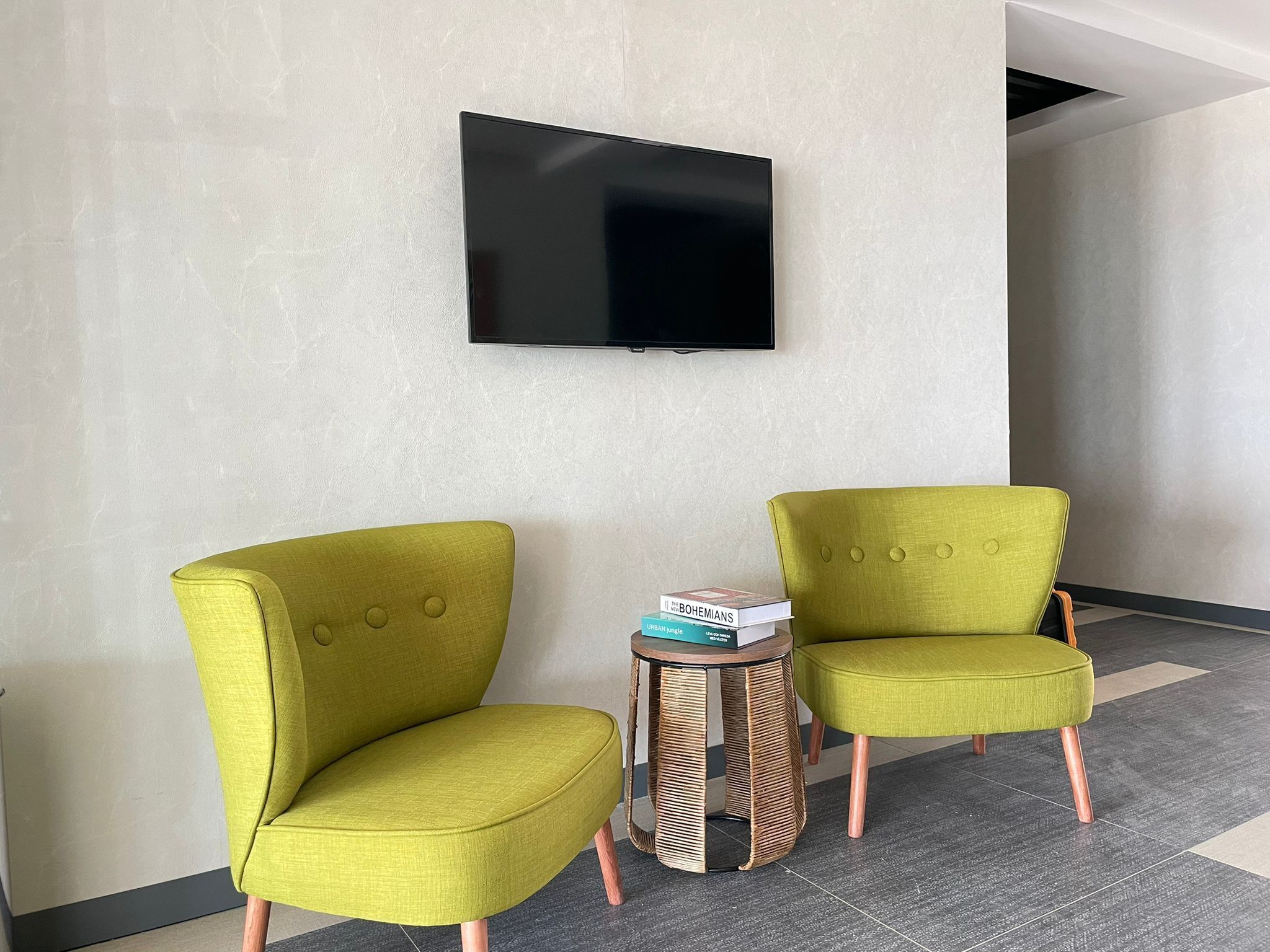 Quarto com duas poltronas verdes estofadas, uma mesinha redonda com base trançada e uma televisão de tela plana fixada na parede.