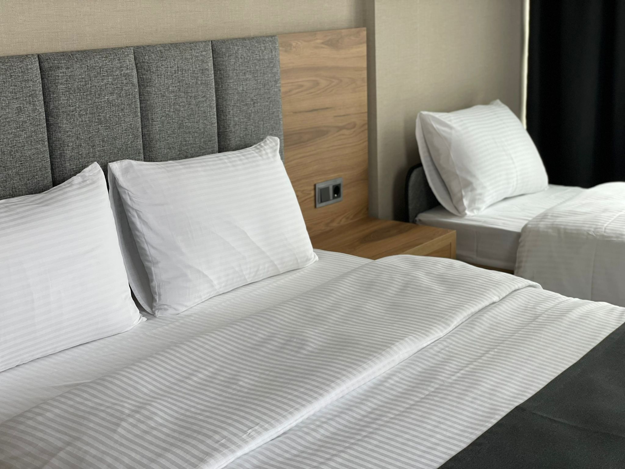 A hotel room with two beds, each with white linens and pillows. The headboard is grey with a wooden panel, and there is a nightstand with a lamp between the beds.