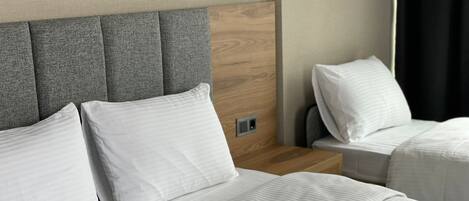 A hotel room with two beds, each with white linens and pillows. The headboard is grey with a wooden panel, and there is a nightstand with a lamp between the beds.