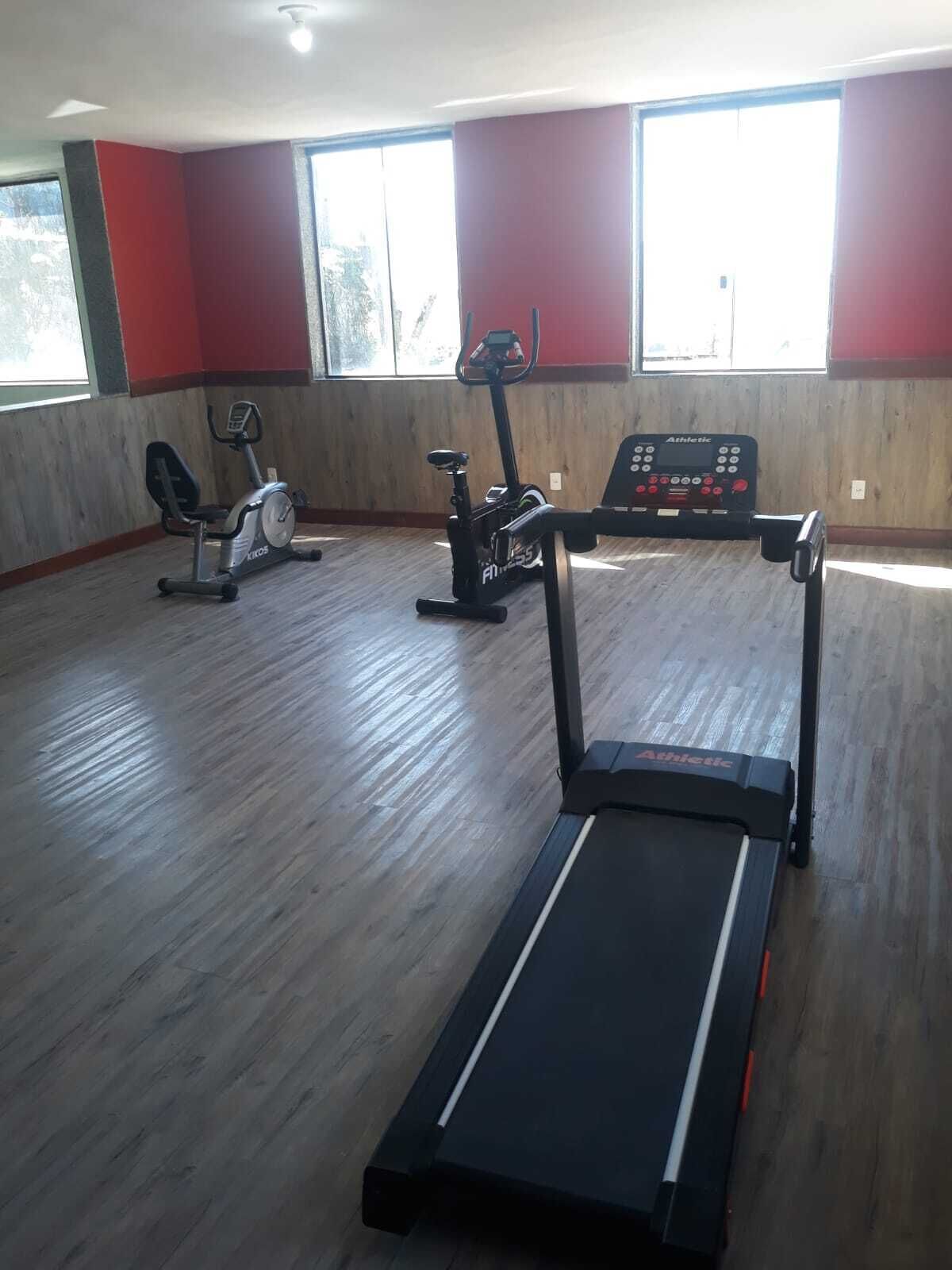 A gym with a treadmill, stationary bikes, and an elliptical in a room with wooden flooring and large windows.