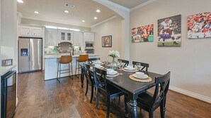 Un comedor con una mesa de madera dispuesta para seis personas, rodeada de sillas. Se observa una cocina al fondo, con armarios blancos y electrodomésticos de acero inoxidable.