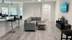 Een moderne woonkamer met een grijze modulaire bank, een houten salontafel en een televisie die aan de muur is bevestigd. Er is een keuken met barkrukken en een eettafel te zien.