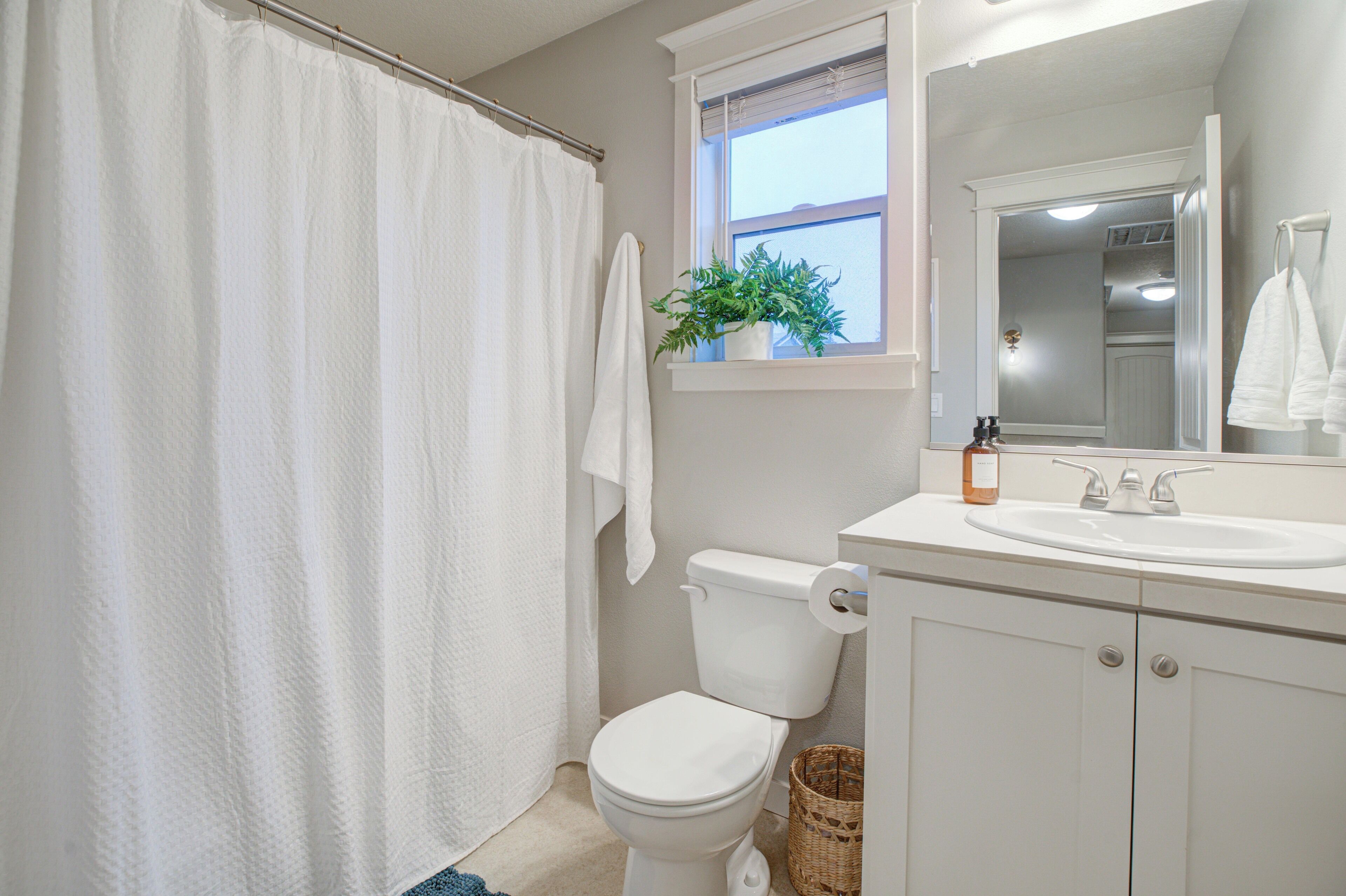 Une salle de bain avec des toilettes blanches, un lavabo blanc, une fenêtre avec une plante et un rideau de douche.