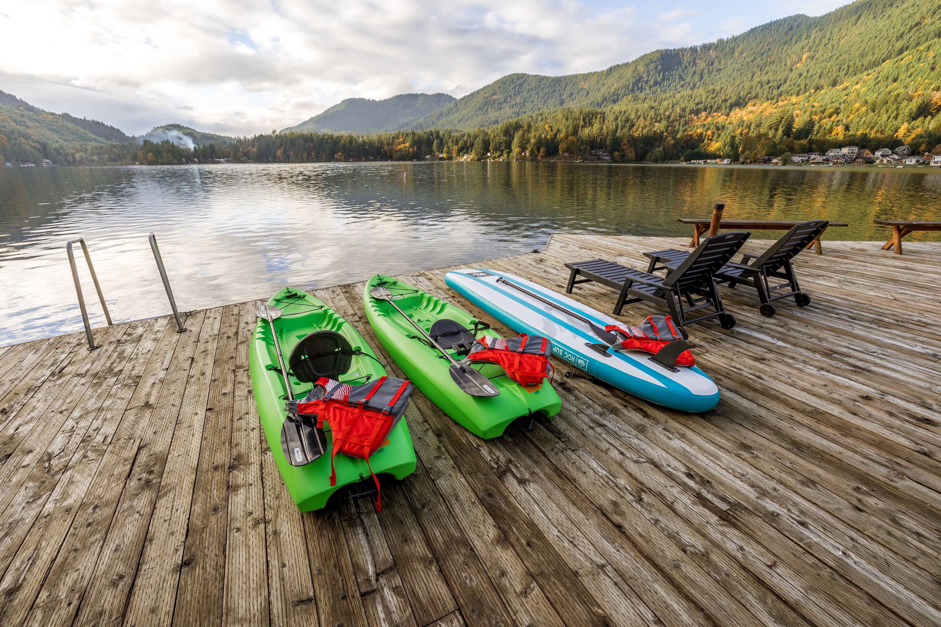 A wooden dock with kayaks and paddleboards, overlooking a calm lake and forested hills.