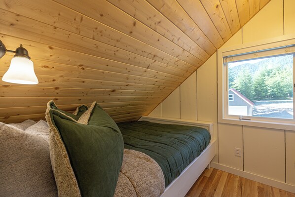 Ein gemütliches Schlafzimmer unter der Schräge mit einem Bett mit grüner Bettwäsche und einem Fenster mit Blick auf ein Haus und Bäume.