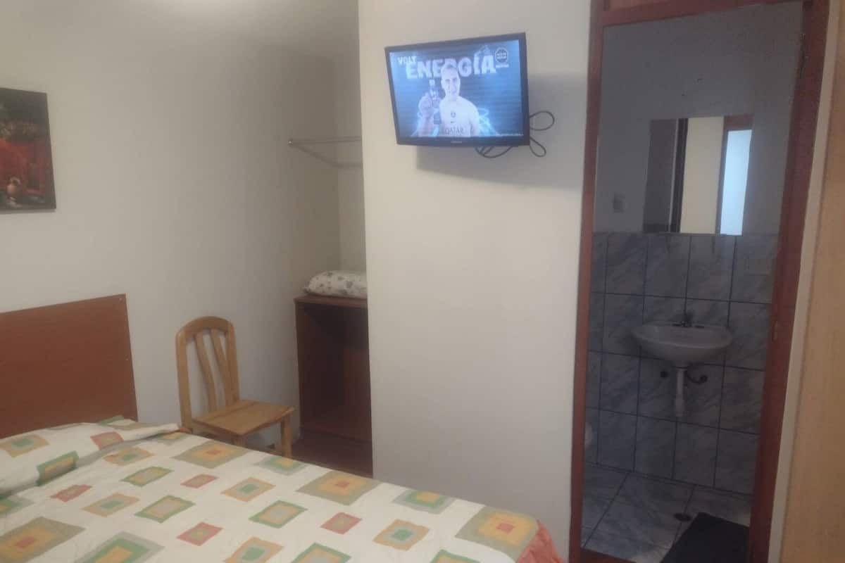 Ein Schlafzimmer mit Bett, Stuhl, Fernseher und einem durchgehenden Türrahmen erkennbaren Badezimmer.