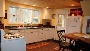 A kitchen with white cabinets, a stove, a sink, and a dining area with a table and chairs. - SUGAR MAPLE CHALET (Dansville)