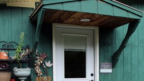 A green wooden shed with a white door, a welcome sign, and potted plants. - SUGAR MAPLE CHALET (Dansville)