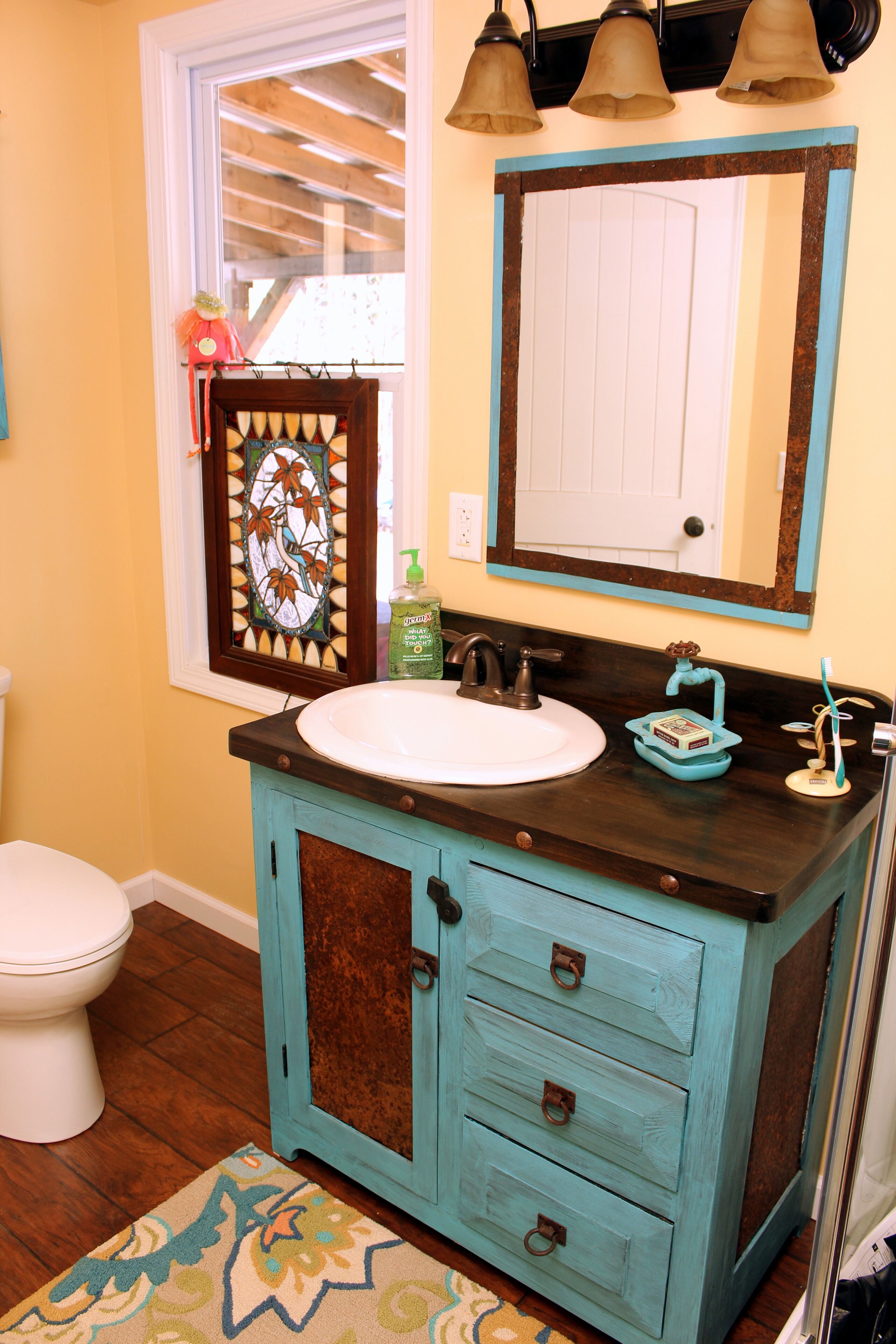 A bathroom with a white toilet, a turquoise vanity with drawers, a mirror, and a window with blinds.