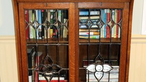 A wooden bookcase with glass doors and metal grilles, containing various books and framed photographs. - SUGAR MAPLE CHALET (Dansville)