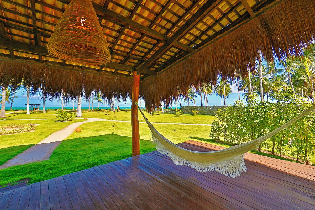 A hammock under a thatched roof, a wooden deck, and a view of the ocean and palm trees.