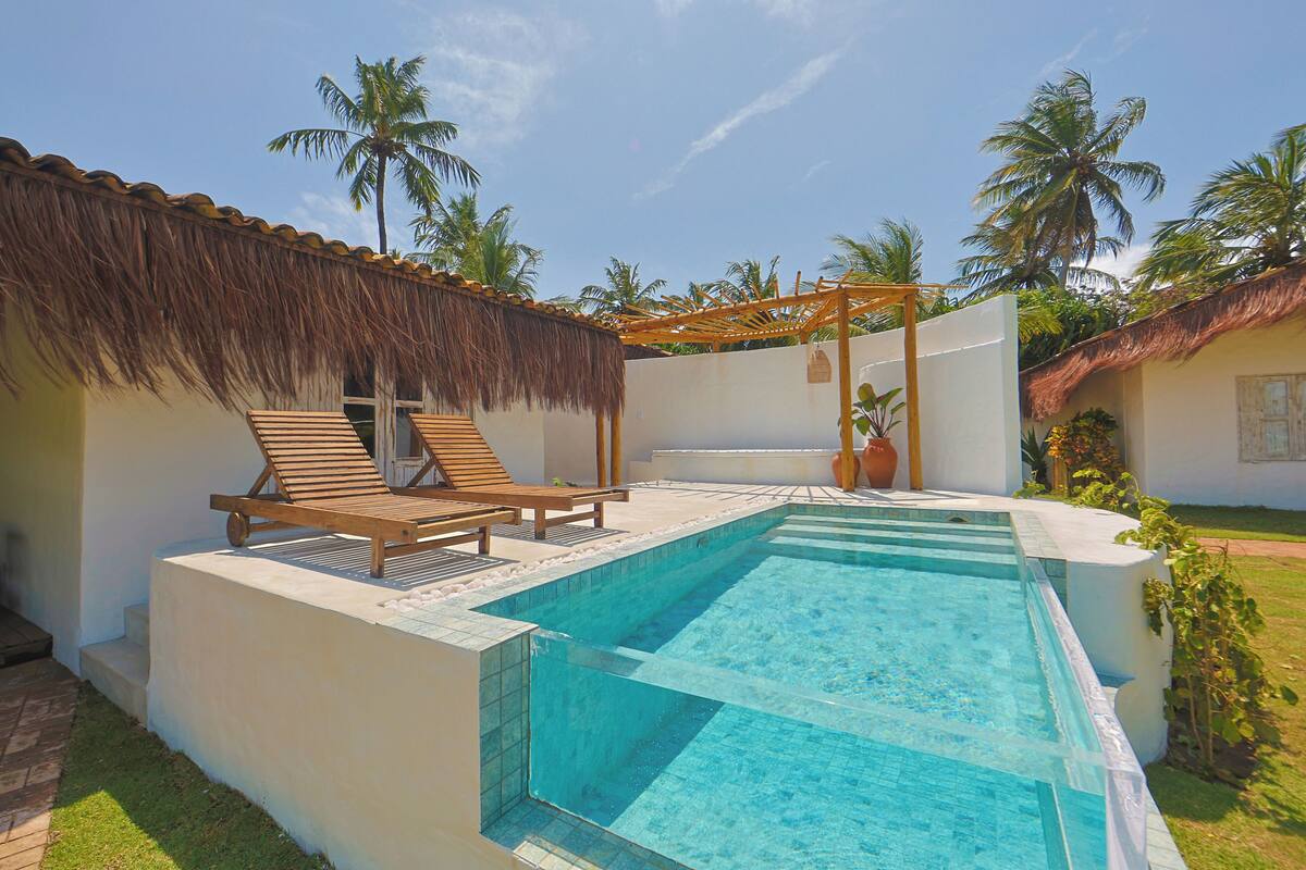 A swimming pool with lounge chairs and a pergola, surrounded by palm trees and a thatched roof structure.