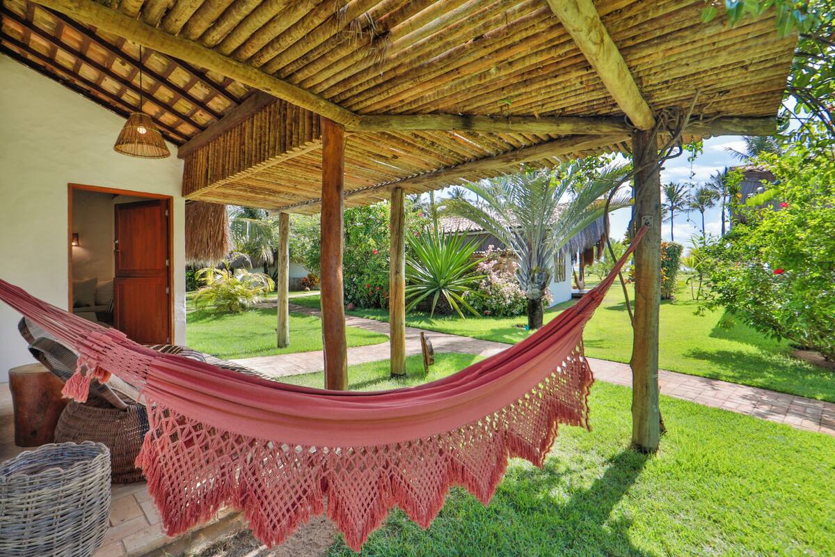 A hammock under a wooden pergola in a lush garden with a pathway and a building entrance.