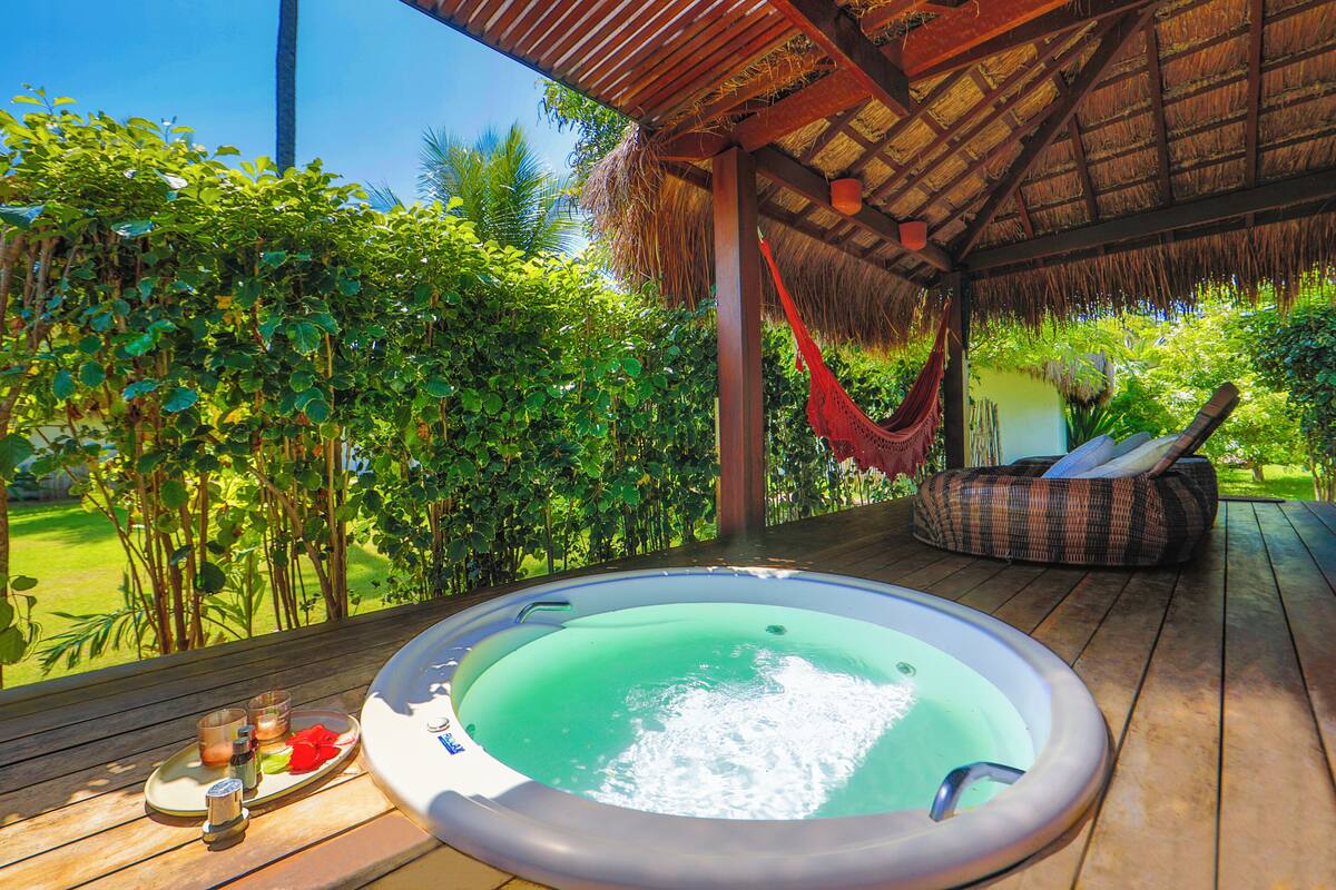 A hot tub on a wooden deck with a thatched roof, surrounded by lush greenery.