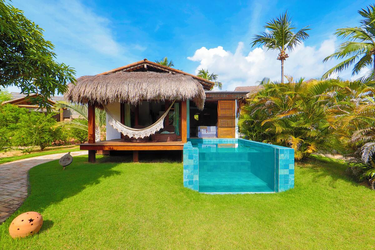 A thatched-roof bungalow with a swimming pool, surrounded by lush greenery and palm trees.