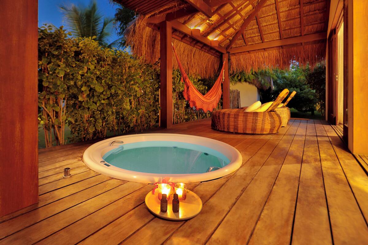A wooden deck with a hot tub, surrounded by lush greenery and a thatched-roof structure.