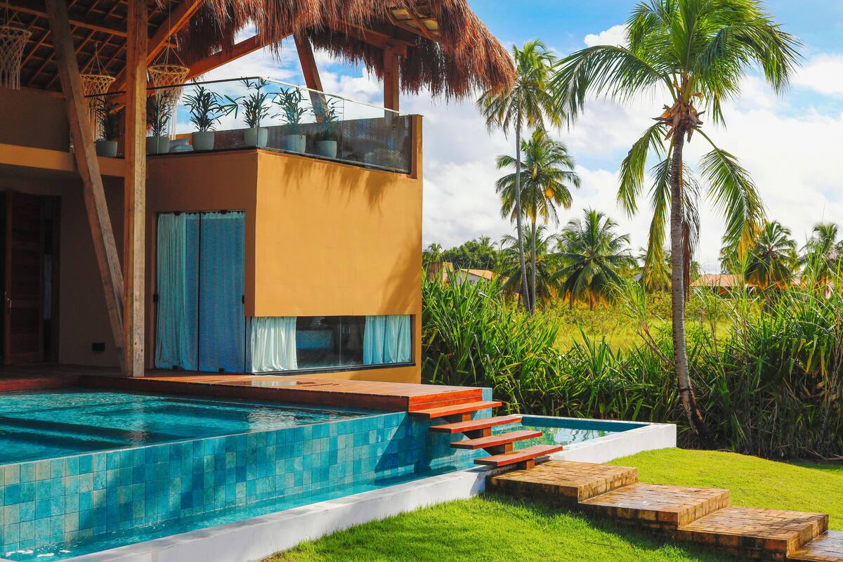 A modern house with a swimming pool, a thatched roof, and palm trees in the background.