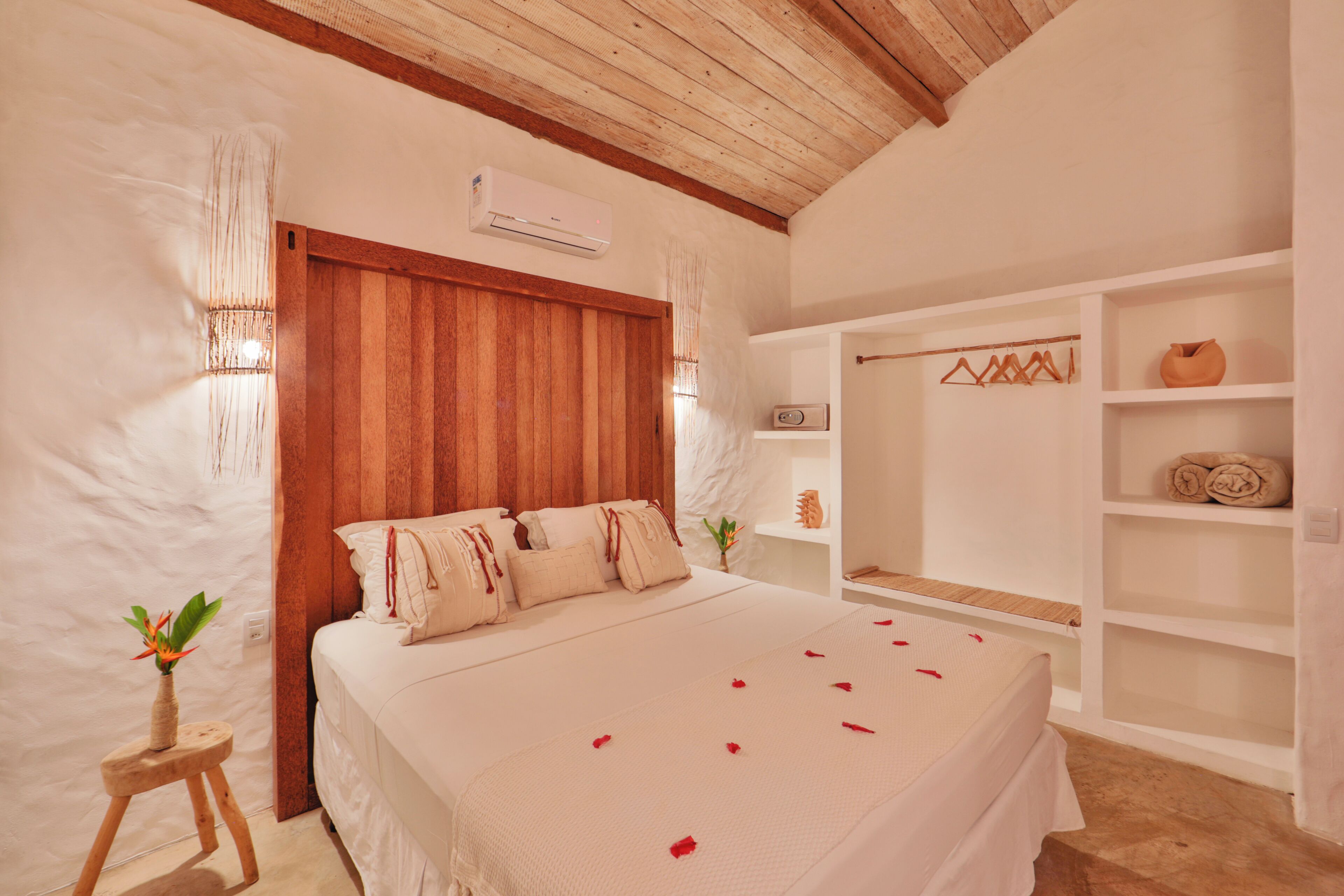 A bedroom with a wooden ceiling, a bed with white bedding, a small wooden stool with a plant, and a white shelf with towels and a vase.