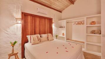 A bedroom with a wooden ceiling, a bed with white bedding, a small wooden stool with a plant, and a white shelf with towels and a vase.