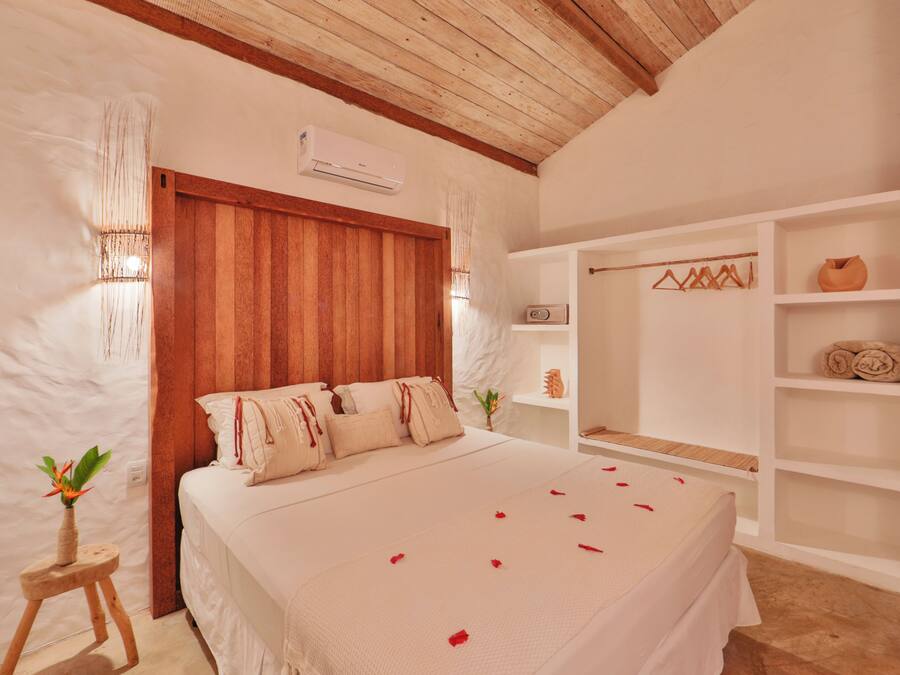 A bedroom with a wooden ceiling, a bed with white bedding, a small wooden stool with a plant, and a white shelf with towels and a vase.