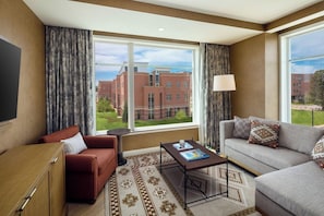 A hotel room with a large window, a sofa, a chair, a coffee table, and a TV. - The Benson Hotel and Faculty Club (Aurora)