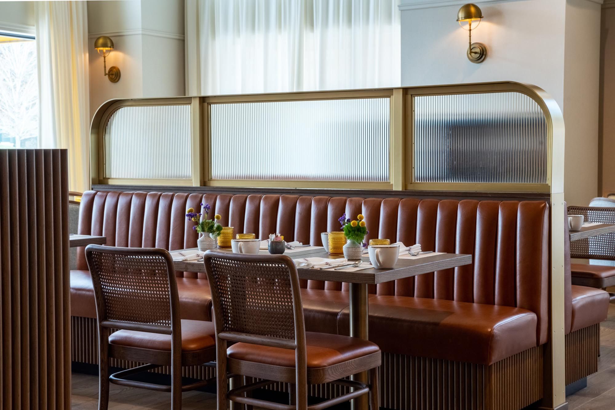 L’intérieur d’un restaurant avec un coin salon composé de banquettes, de chaises en bois et d’une table dressée avec des assiettes, des tasses et un vase de fleurs.