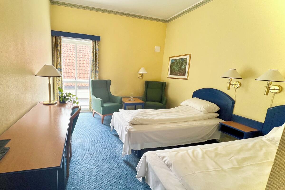 Une chambre d’hôtel avec un lit, deux chaises, un bureau avec une télévision et une fenêtre avec des rideaux.