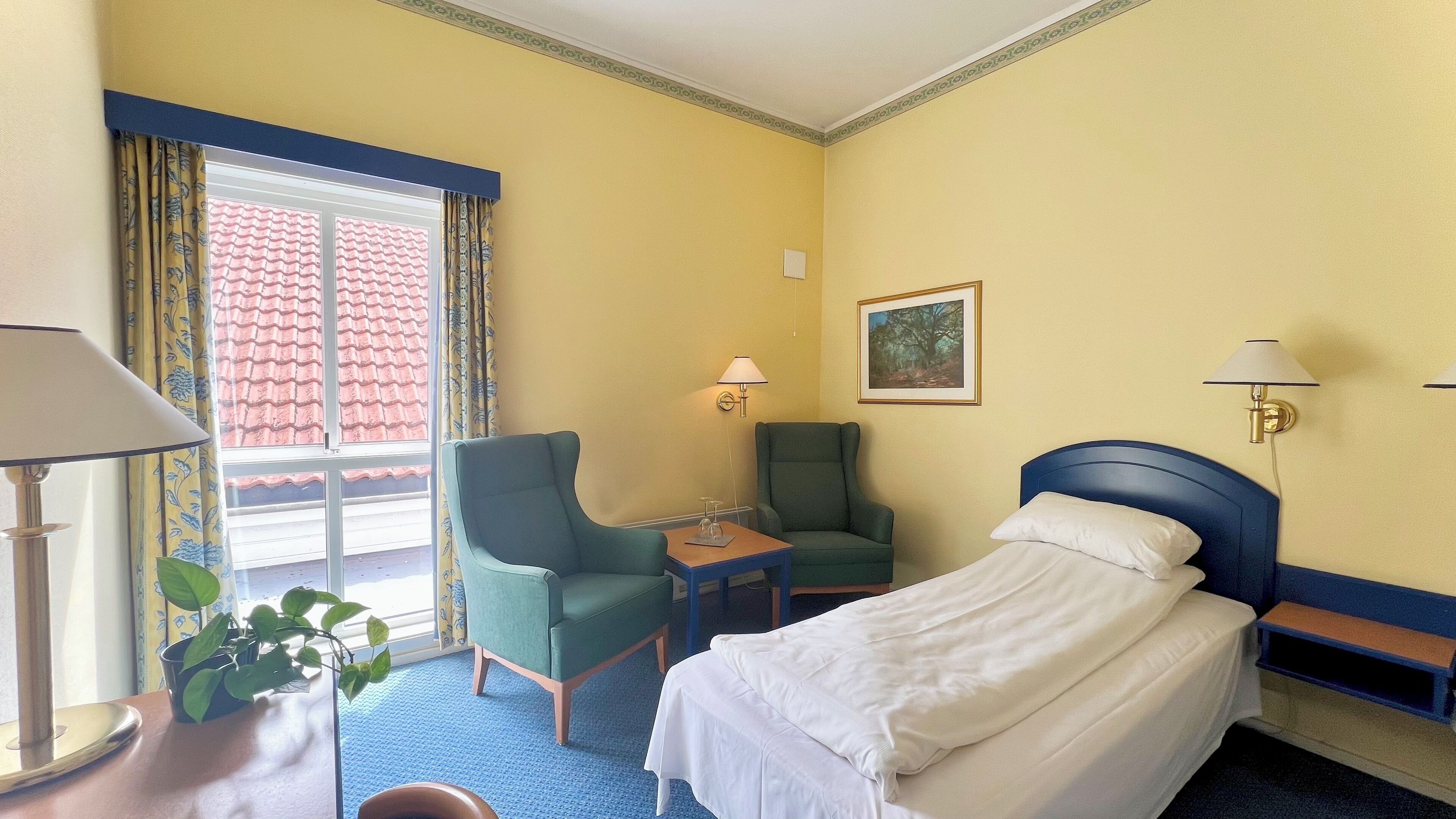 A hotel room with a bed, a desk, a chair, a lamp, a window with curtains, and a painting on the wall.