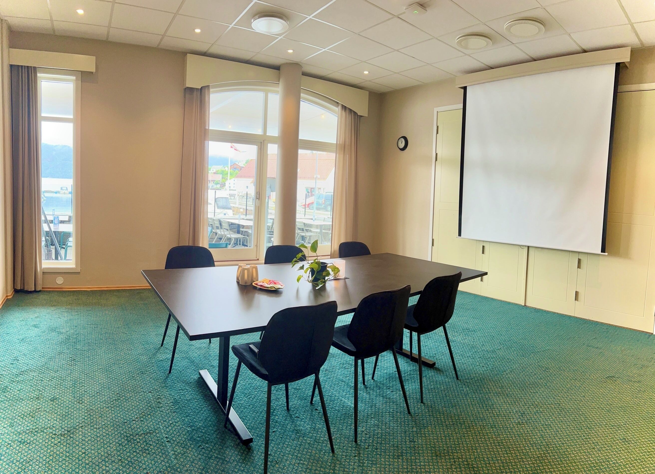 A conference room with a large table, four chairs, a projector screen, and a view of the city through large windows.