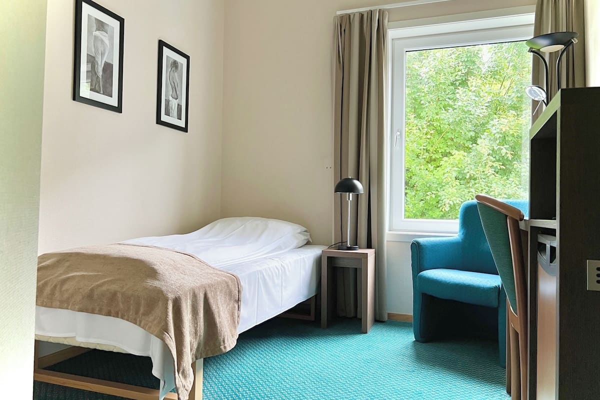 Une chambre d’hôtel avec un lit, une chaise, un bureau, une lampe et une fenêtre avec des rideaux.