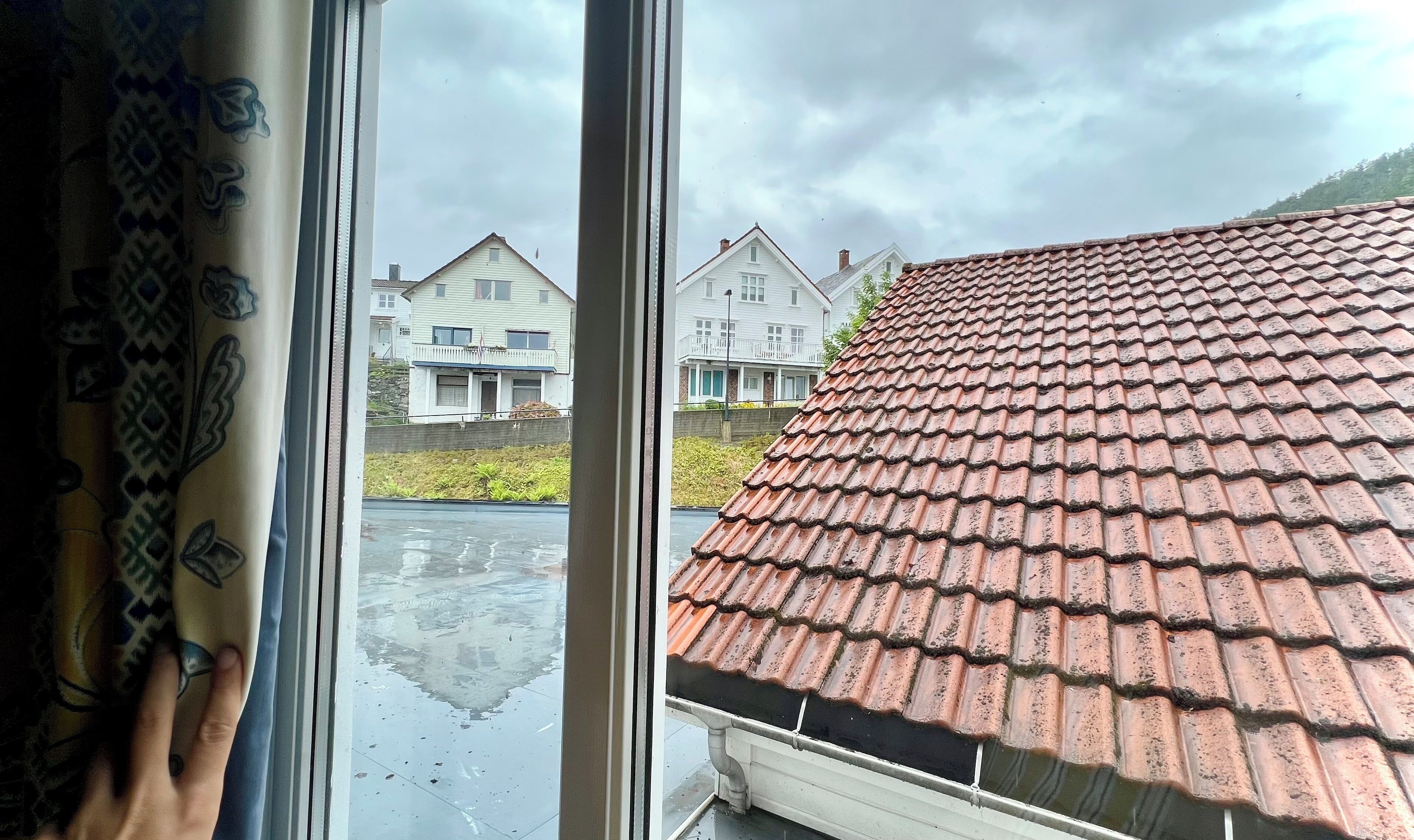 Vista a través de una ventana que muestra una zona residencial con tejados rojos y un edificio blanco con balcón.