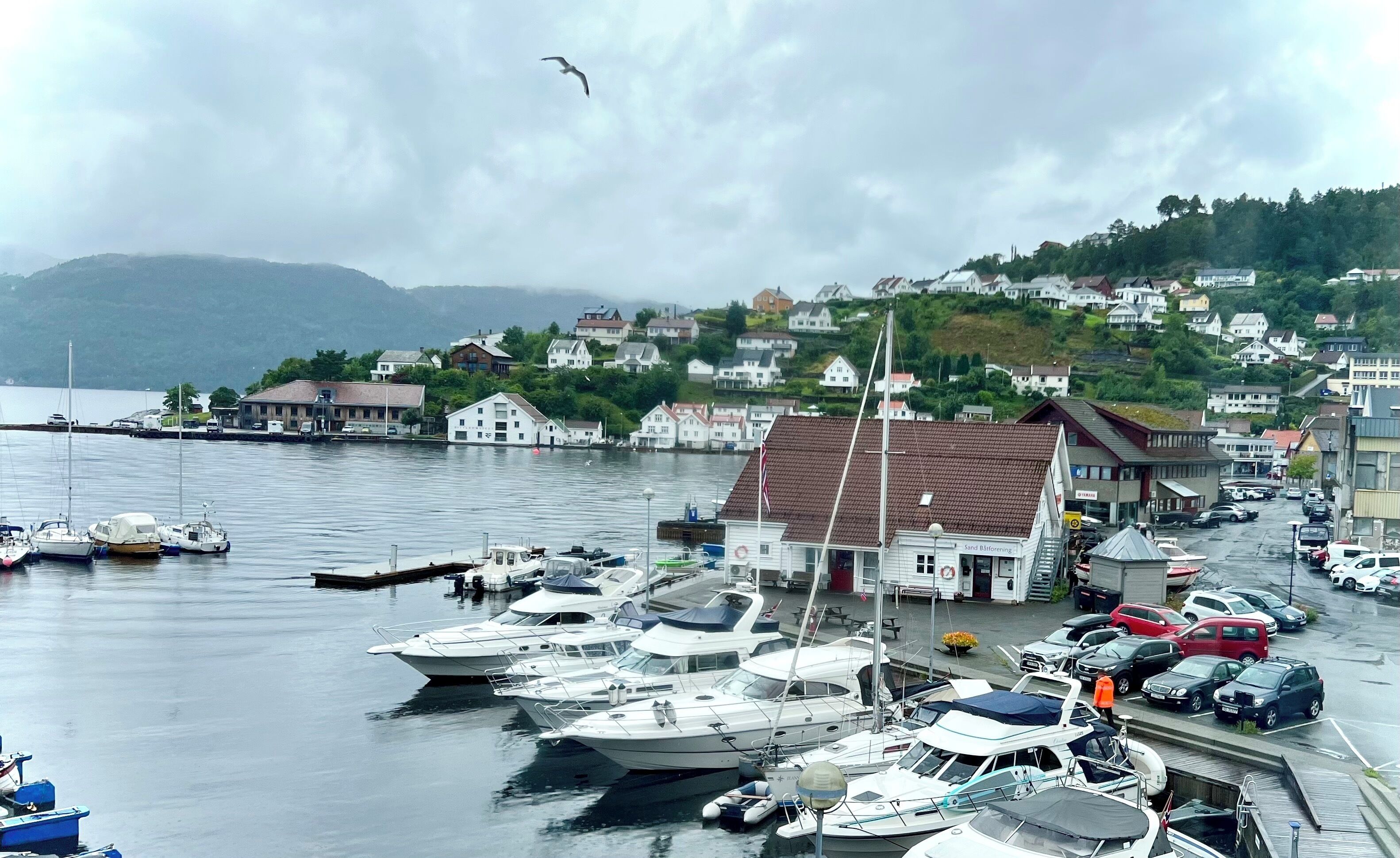 Un port de plaisance où sont amarrés plusieurs bateaux, un bâtiment sur lequel on peut lire l’inscription « Sand Børøying », et une colline avec des maisons en arrière-plan.