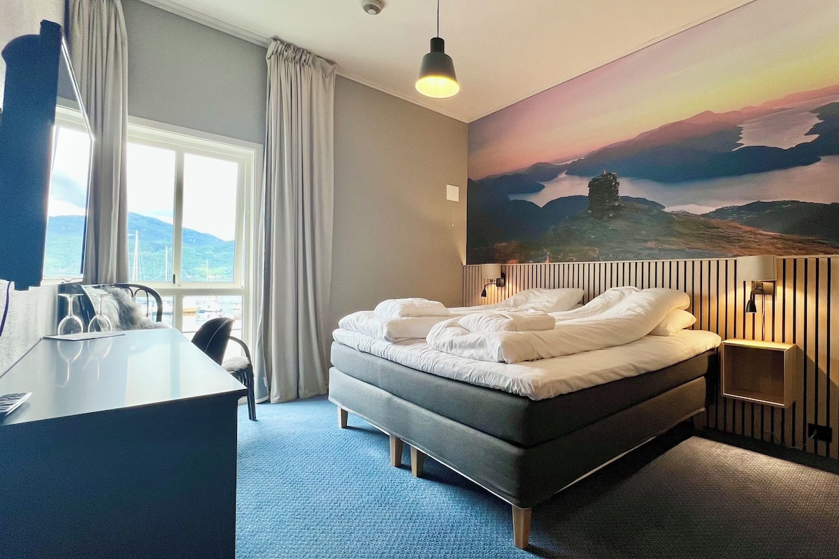 Une chambre d’hôtel comprenant un lit, un bureau, une chaise, une fenêtre avec des rideaux, un tableau représentant un paysage et une lampe.
