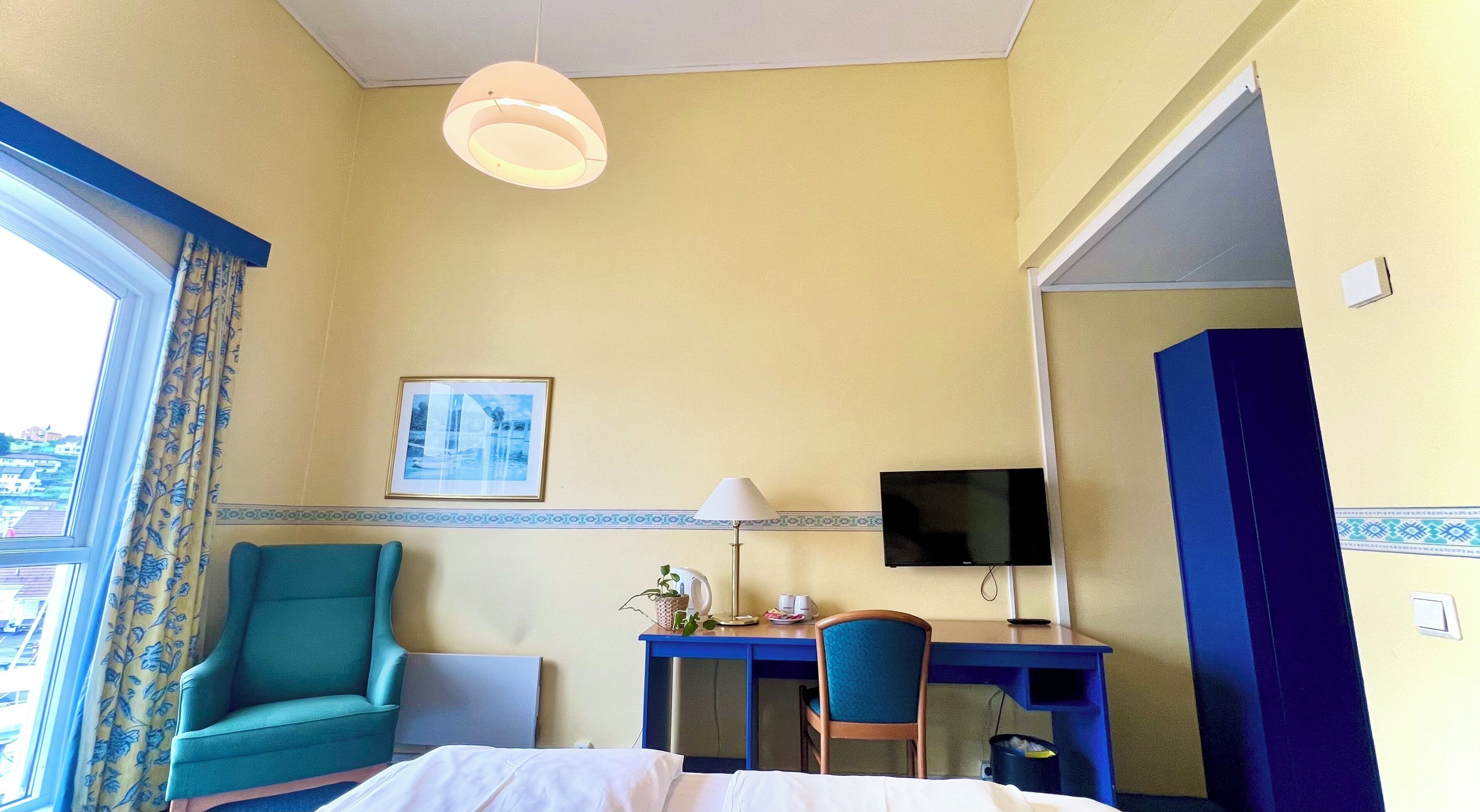 Ett hotellrum med en säng, ett skrivbord med en stol, en tv och ett fönster med gardiner.