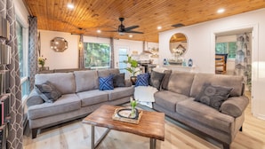 A modern living room with a wooden ceiling, a sectional sofa, a coffee table, and a kitchen in the background.