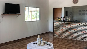 A spacious living room with a large TV, a round coffee table with decorative items, a woven rug, and a brick accent wall. - Pousada Mar das Gaivotas (Cajueiro da Praia)