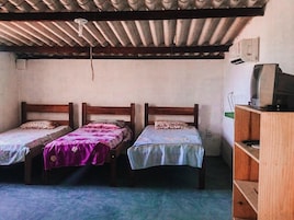 Quarto pequeno com duas camas de madeira, uma estante com uma televisão e teto de metal corrugado.