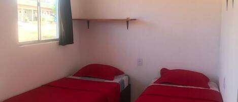 A room with two single beds, a window with curtains, an air conditioning unit, and a small shelf.